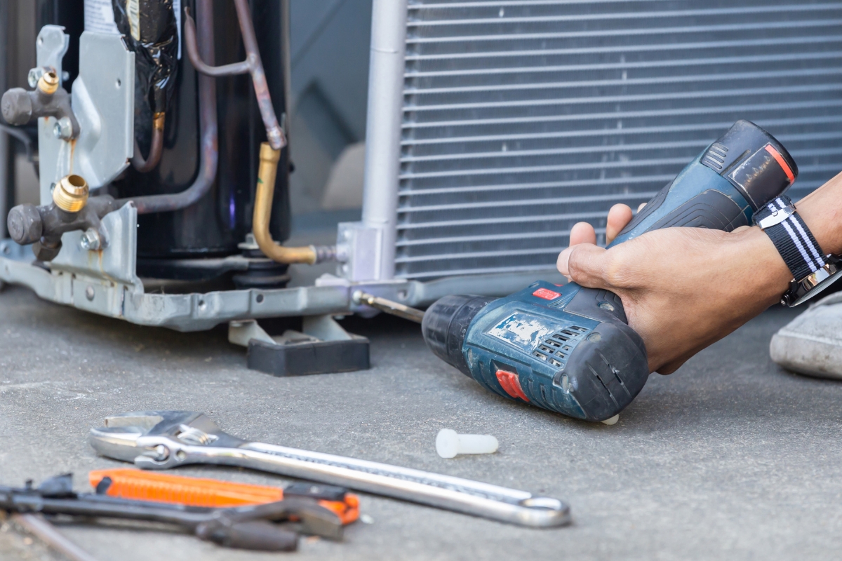 An HVAC technician using a cordless drill to service an air conditioning condenser unit, with hand tools laid out nearby